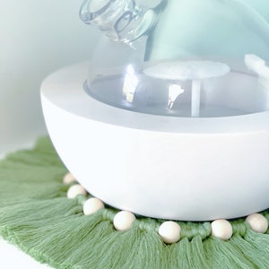May include: A white, spherical essential oil diffuser with a clear glass top. It sits on a green macrame coaster with wooden beads. The diffuser has a modern, minimalist design, perfect for aromatherapy.