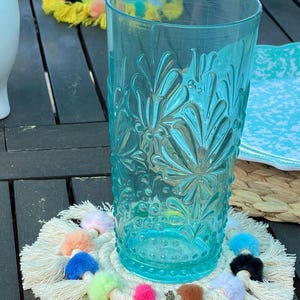 May include: A white crocheted coaster with colorful pom-poms and a small llama charm. The coaster is placed under a blue glass with a floral pattern.