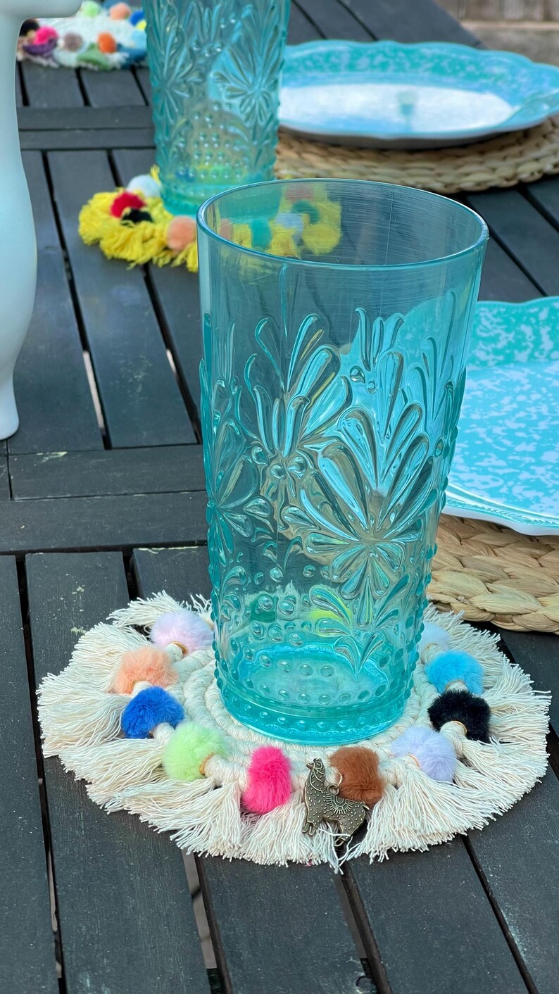 May include: A white crocheted coaster with colorful pom-poms and a small llama charm. The coaster is placed under a blue glass with a floral pattern.