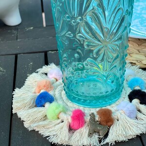 May include: A white crocheted coaster with colorful pom-poms and tassels. A small gold llama charm is attached to the coaster. A blue glass with a floral pattern sits on top of the coaster.