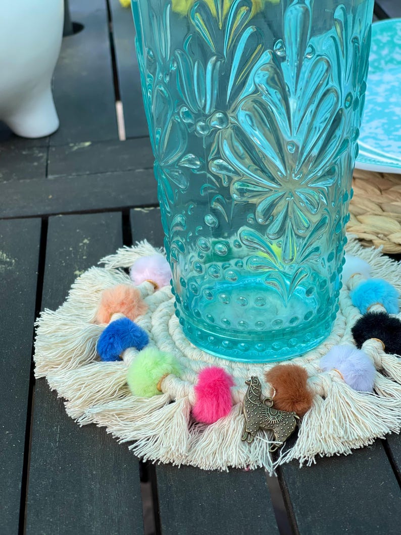May include: A white crocheted coaster with colorful pom-poms and tassels. A small gold llama charm is attached to the coaster. A blue glass with a floral pattern sits on top of the coaster.