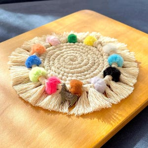 May include: A round, beige, crocheted coaster with a fringe border. The coaster is decorated with colorful pom-poms and a small metal llama charm.
