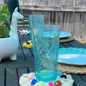 May include: Two blue glass tumblers with a textured design, one on a white crocheted coaster with colorful pom-poms and a gold llama charm.