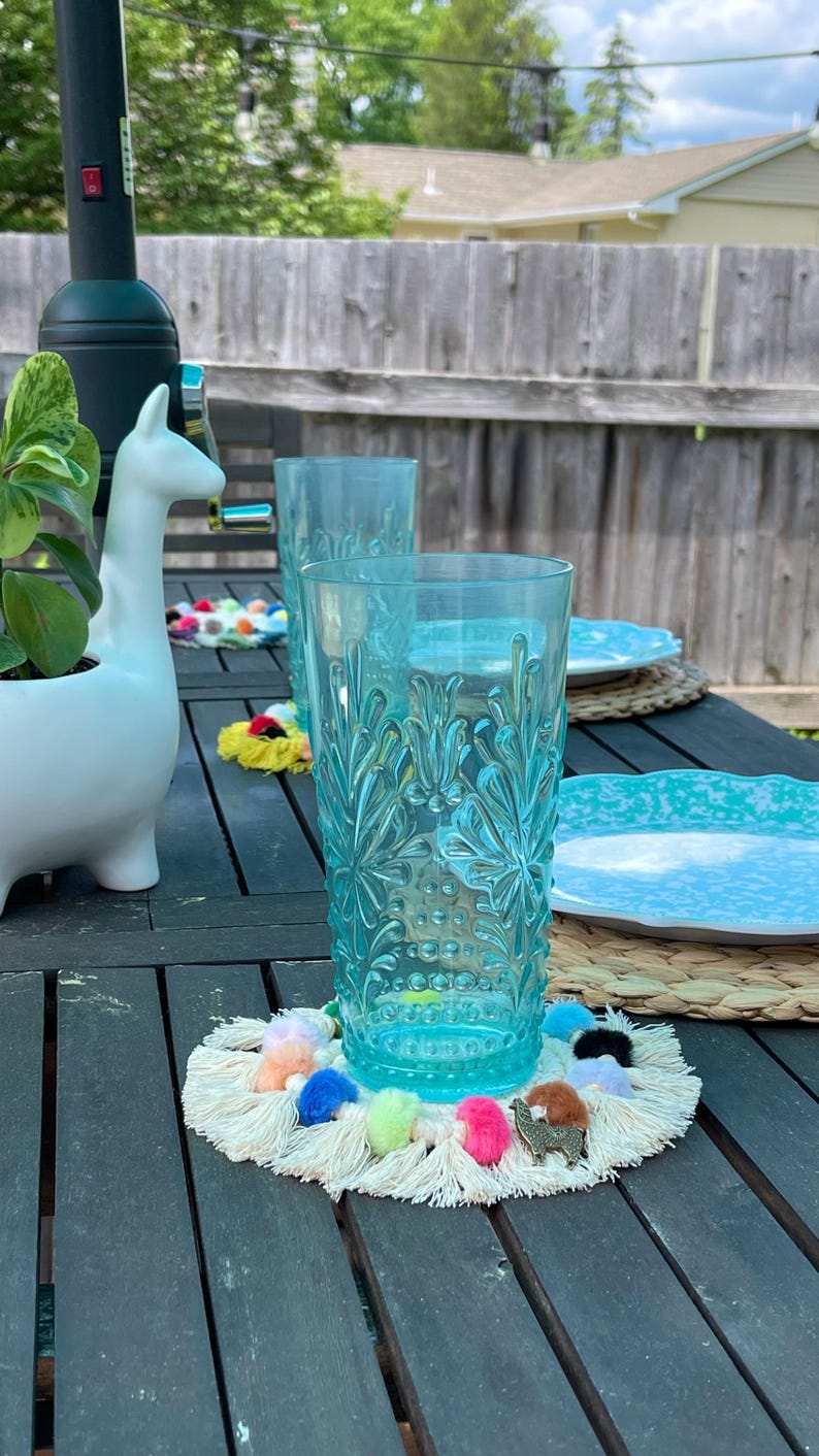 May include: Two blue glass tumblers with a textured design, one on a white crocheted coaster with colorful pom-poms and a gold llama charm.