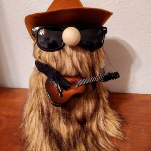 May include: A whimsical, furry creature with a brown cowboy hat, sunglasses, and a wooden nose. It wears a black scarf and holds a miniature acoustic guitar. The creature's fur is a mix of brown and tan.