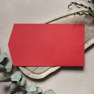 May include: A red envelope with a flap on the left side. The envelope is lying on a wooden surface.