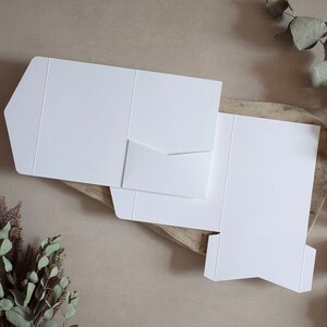 May include: White cardstock pocket folders, partially assembled, with a small pocket on the left side. The folders are on a wooden surface with greenery in the background. The folders are blank and ready for customization.