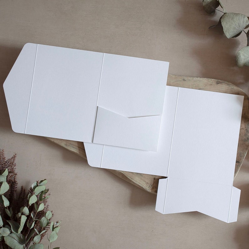 May include: White cardstock pocket folders, partially assembled, with a small pocket on the left side. The folders are on a wooden surface with greenery in the background. The folders are blank and ready for customization.