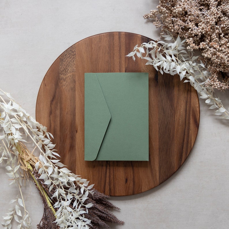 May include: A green envelope on a wooden surface with white flowers and dried branches.
