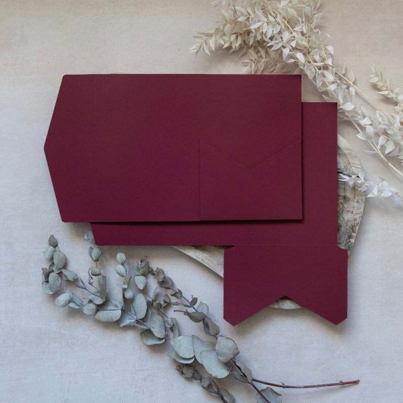 Puede incluir: Una tarjeta de color burdeos con un dise&ntilde;o plegado. La tarjeta est&aacute; sobre una superficie de madera con flores secas.