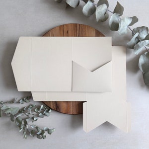 Puede incluir: Un conjunto de tres sobres de papel de color blanco roto con un dise&ntilde;o &uacute;nico de pliegue. Los sobres est&aacute;n apilados sobre una superficie de madera.