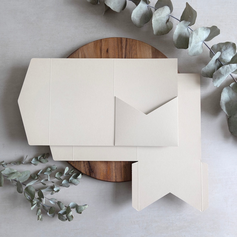 Puede incluir: Un conjunto de tres sobres de papel de color blanco roto con un dise&ntilde;o &uacute;nico de pliegue. Los sobres est&aacute;n apilados sobre una superficie de madera.