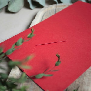 May include: A red envelope with a white flap. The envelope is lying on a wooden surface.