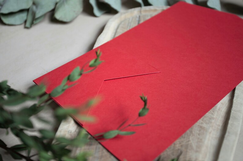 May include: A red envelope with a white flap. The envelope is lying on a wooden surface.