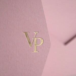 May include: A close-up of a pink envelope with a gold foil stamp of the letters "VP". The envelope has a textured surface and a unique angled flap design. The background is a soft, blurred white.