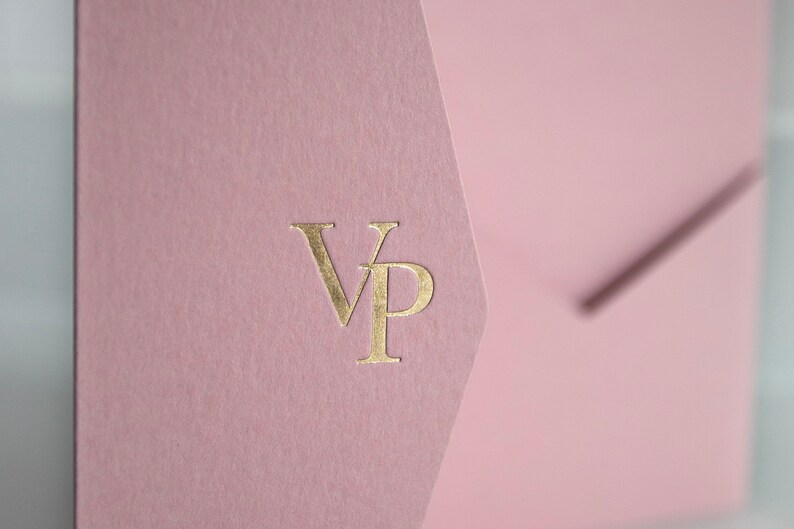 May include: A close-up of a pink envelope with a gold foil stamp of the letters "VP". The envelope has a textured surface and a unique angled flap design. The background is a soft, blurred white.