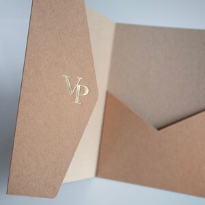 May include: A tan presentation folder with a pocket, featuring a gold foil stamped "VP" monogram. The folder is open, revealing a lighter tan interior. The paper has a textured finish.