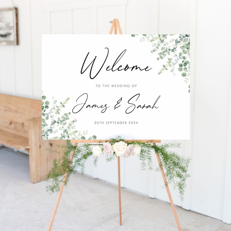 May include: White wedding welcome sign with green eucalyptus leaves and the text "Welcome to the wedding of James & Sarah 20th September 2024".