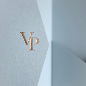 May include: Close-up of a light blue paper with a gold foil stamped "VP" monogram. The paper has a textured finish and is folded, revealing a glimpse of a light blue background.