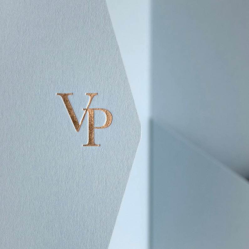 May include: Close-up of a light blue paper with a gold foil stamped "VP" monogram. The paper has a textured finish and is folded, revealing a glimpse of a light blue background.