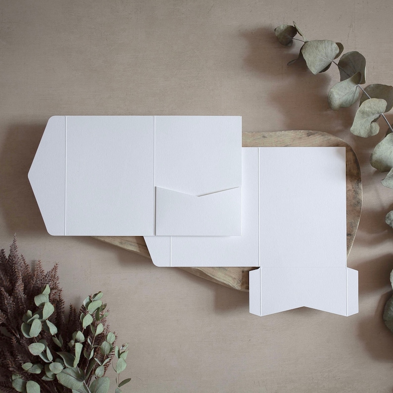 May include: White invitation card templates in various stages of assembly, with a pocket and flap design. The cards are arranged on a wooden surface, with sprigs of eucalyptus and dried flowers adding a natural touch.