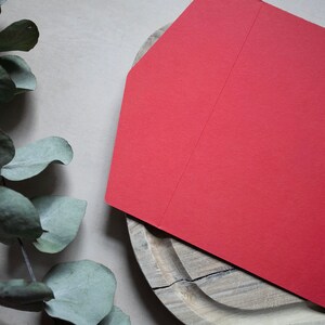May include: A red envelope with a folded flap on a wooden surface. The envelope is blank and has a textured surface.