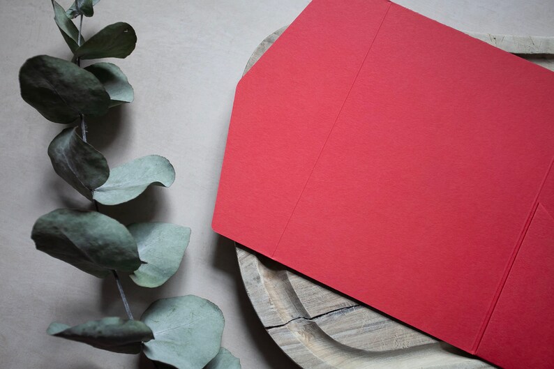 May include: A red envelope with a folded flap on a wooden surface. The envelope is blank and has a textured surface.