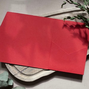 May include: A red envelope with a flap on the left side. The envelope is lying on a wooden surface with green leaves in the background.