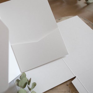 May include: A close-up of several white paper pocket folders, with a textured finish. The folders are arranged at different angles, with one open to reveal the pocket. A sprig of green leaves is in the lower left corner.