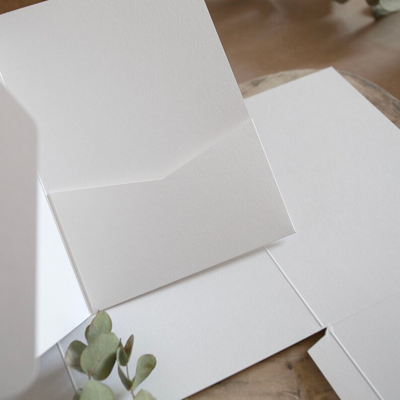 May include: A close-up of several white paper pocket folders, with a textured finish. The folders are arranged at different angles, with one open to reveal the pocket. A sprig of green leaves is in the lower left corner.