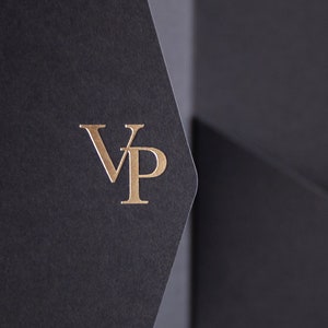 May include: Close-up of a black presentation folder with a gold foil stamped "VP" logo. The folder is made of textured paper and has a pocket. The background is a soft, neutral color.