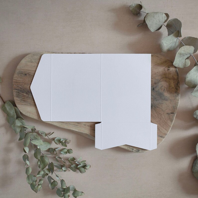 May include: A white, unfolded invitation envelope with a unique flap design, resting on a wooden oval board. Eucalyptus branches frame the scene, creating a natural, minimalist aesthetic. The envelope is blank, ready for customization.
