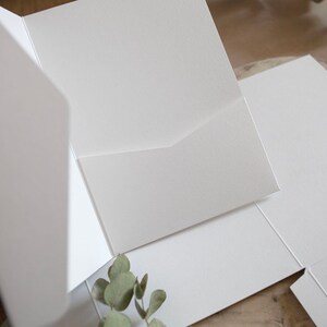 May include: A close-up of several white, blank pocket folders, with a textured surface. The folders are open and arranged on a wooden surface, with a sprig of greenery in the corner.