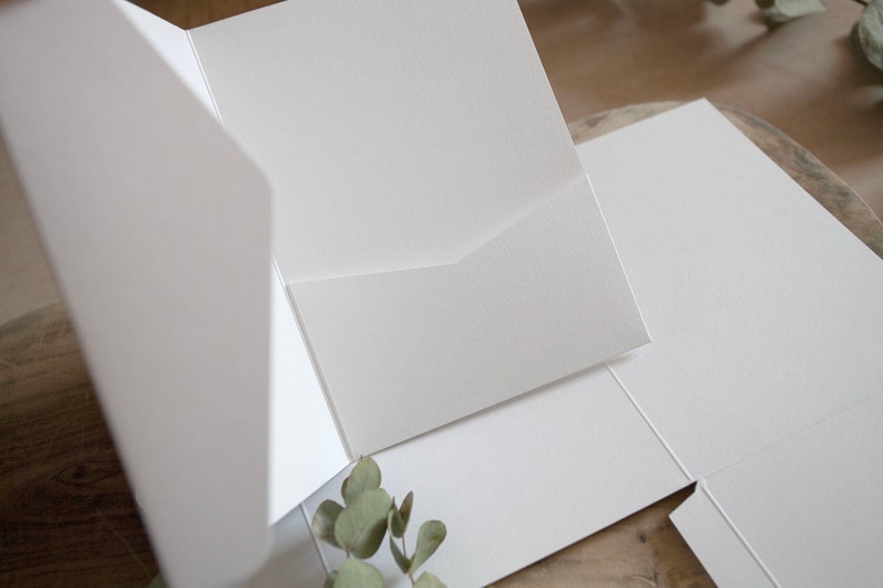 May include: A close-up of several white, blank pocket folders, with a textured surface. The folders are open and arranged on a wooden surface, with a sprig of greenery in the corner.