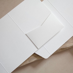 May include: A close-up of a white stationery set, including a folded card and an envelope. The set is made of textured paper and is arranged on a light-colored surface. The card is partially open, revealing the inside.