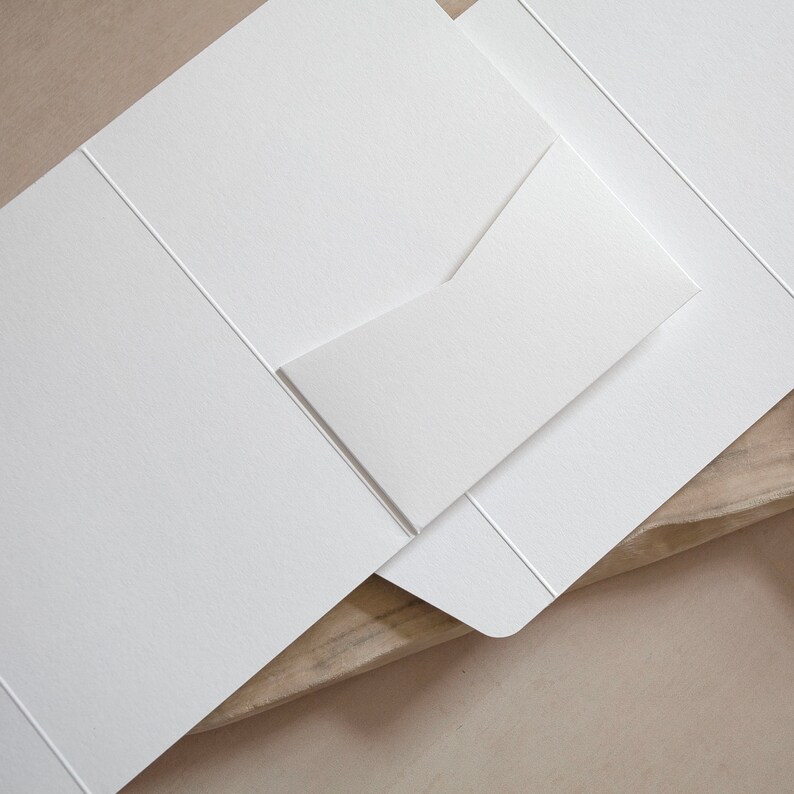 May include: A close-up of a white stationery set, including a folded card and an envelope. The set is made of textured paper and is arranged on a light-colored surface. The card is partially open, revealing the inside.