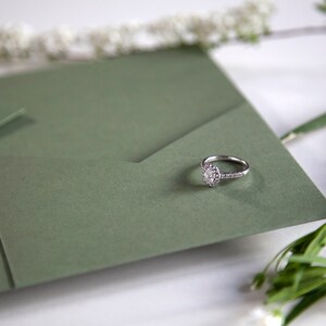 May include: A silver engagement ring with a large, clear diamond set in a halo setting. The ring is resting on a green envelope.