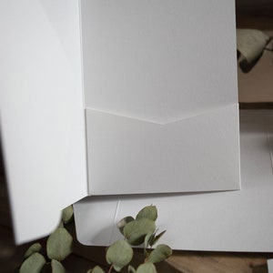 May include: White presentation folders with a pocket. The folders are open, revealing the interior pocket. The image is a close-up, highlighting the texture and design of the folders. Eucalyptus leaves are in the foreground.