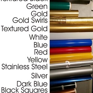 K&ouml;nnte beinhalten: Eine Auswahl an Rollen von Vinyl-Klebefolie in verschiedenen Farben und Texturen, darunter strukturiertes Schwarz, Gr&uuml;n, Gold, Gold mit Swirls, strukturiertes Gold, Wei&szlig;, Blau, Rot, Gelb, Edelstahl, Silber, Dunkelblau, schwarze Quadrate und Bronze.