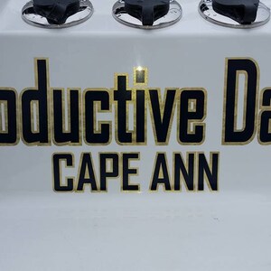 K&ouml;nnte beinhalten: Ein wei&szlig;es Schild mit schwarzem Text, der "Productive Daze" mit goldener Umrandung lautet. Unter dem Text steht "CAPE ANN" in goldenen Buchstaben.