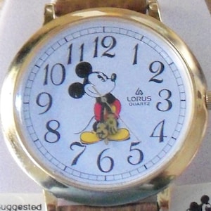 Könnte beinhalten: Eine goldfarbene Mickey Mouse Uhr mit weißem Zifferblatt und schwarzen Zeigern. Die Uhr hat ein braunes Lederarmband und auf dem Zifferblatt steht "Lorus Quartz".
