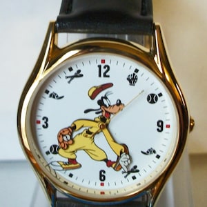 May include: A gold-tone wristwatch with a white face featuring a cartoon image of Goofy playing baseball. The watch has a black leather strap and Roman numeral hour markers.