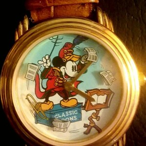 May include: A gold-toned wristwatch with a brown leather band. The watch face features a cartoon image of Mickey Mouse in a conductor's uniform, with musical notes and instruments. The watch has gold hands and a gold bezel.