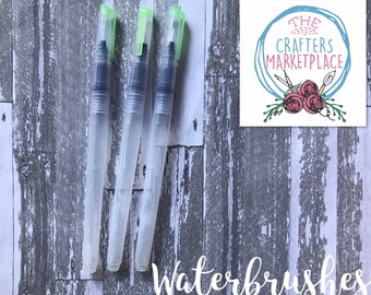 Op de afbeelding: Drie transparante waterpenseelen met groene doppen op een grijze houten achtergrond. De tekst "Waterbrushes" is in witte cursieve letters onder de penseelen geschreven. Een logo voor "The Crafters Marketplace" bevindt zich in de rechterbovenhoek van de afbeelding.