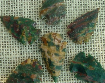 May include: Six arrowheads made of green, brown, and white jasper. The arrowheads are arranged in a star shape on a burlap background.