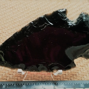 May include: A dark obsidian arrowhead, approximately 6 inches long, with a chipped edge, resting on a woven fabric surface.