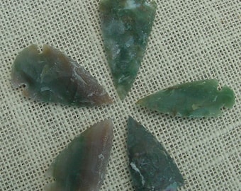 May include: Five green and brown arrowheads made of stone. The arrowheads are arranged in a star shape on a beige fabric background.