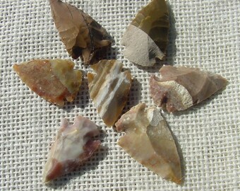 May include: Six arrowheads made of flint, each with a different pattern of brown, white, and gray colors. The arrowheads are arranged in a star shape on a brown fabric background.