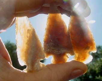 May include: Three arrowheads made of light brown and white stone, held in a hand against a bright blue sky.