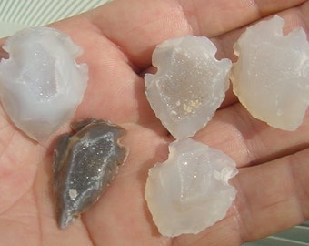 May include: Five arrowhead-shaped stones with a white and gray agate pattern. The stones are translucent and have a natural, rough texture.